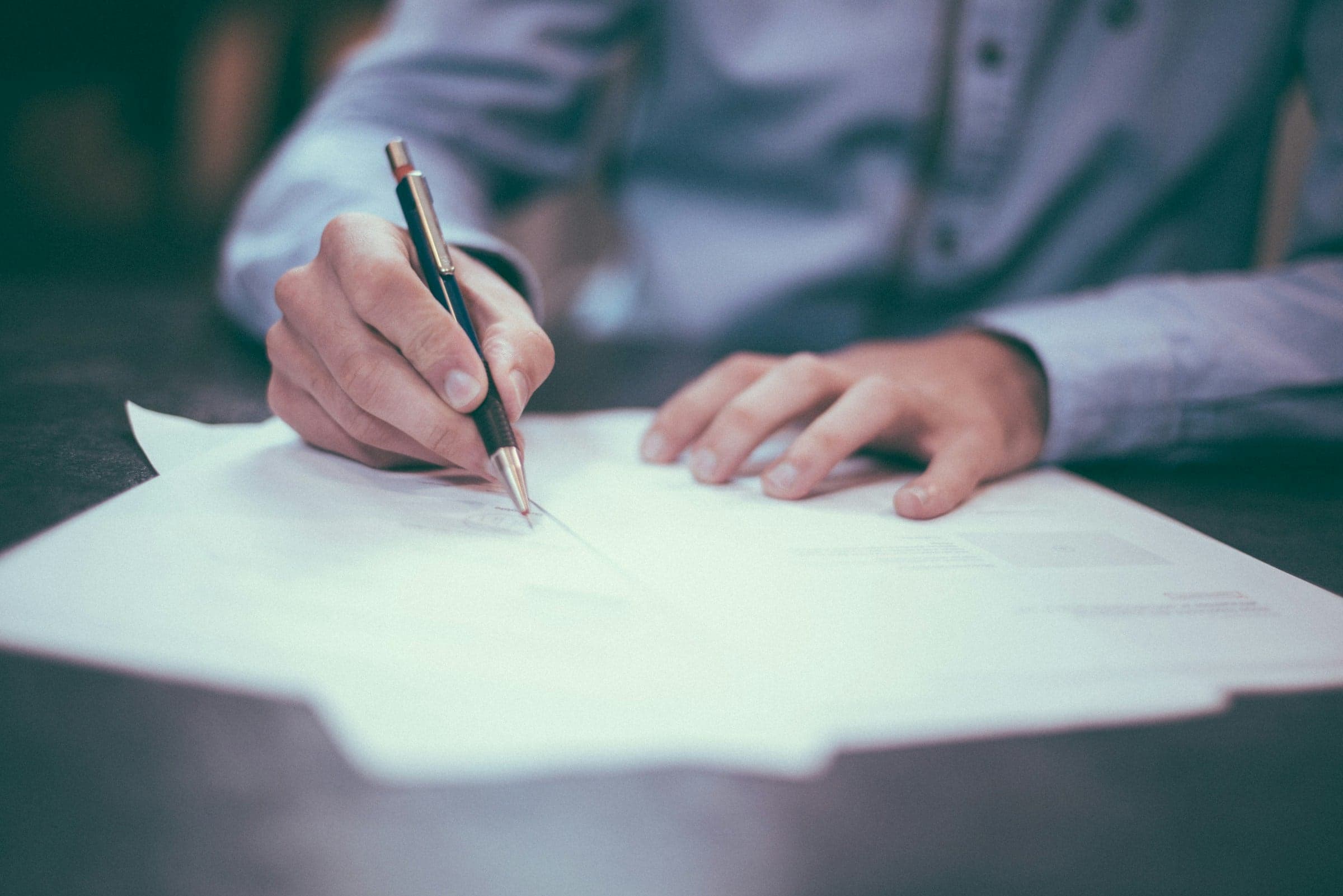 A close-up of a hand signing a document at a desk: the formal signature Pactbound turns into a sealed, witnessed acceptance record.
