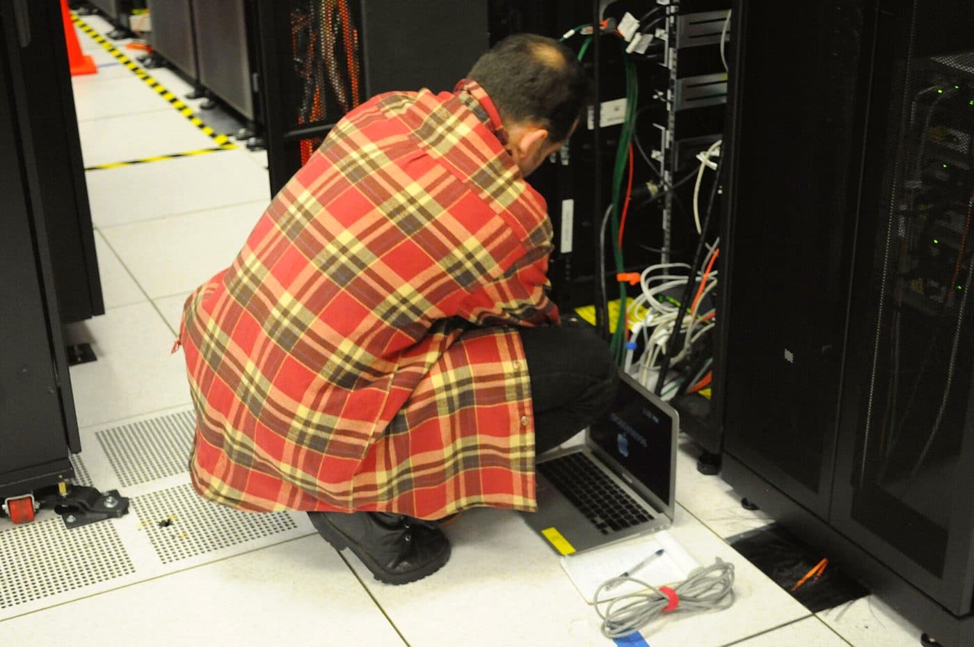 A technician kneeling at an open server cabinet with a laptop, working on the equipment inside.