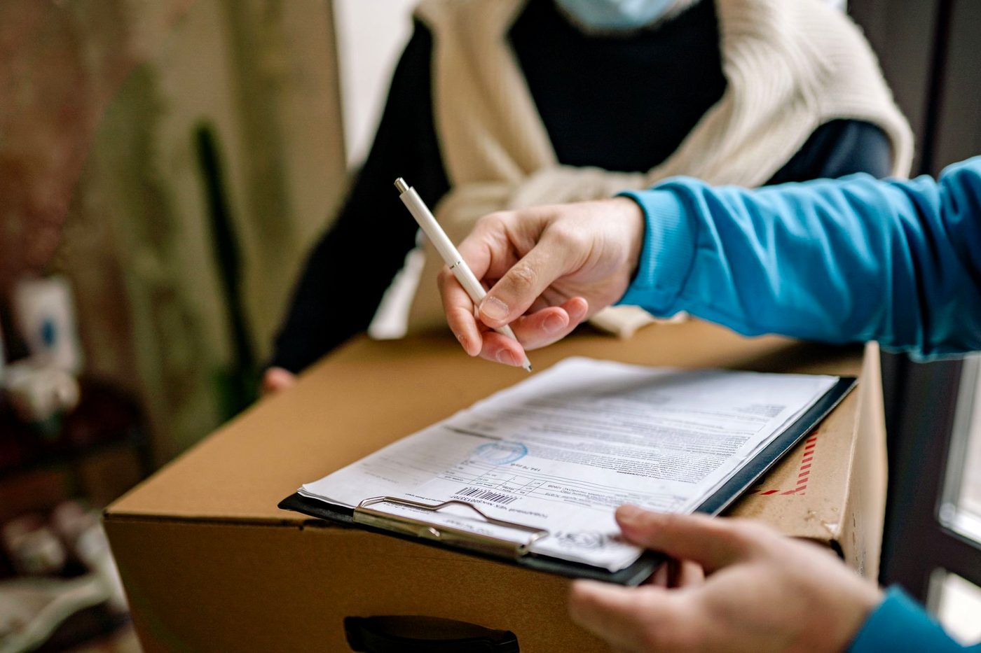 A hand signing a form on a clipboard resting on a cardboard parcel.